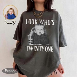 May include: A charcoal gray oversized t-shirt featuring a black and white photograph of a young child with pigtails. The text "LOOK WHO'S" appears above the image, and "TWENTY ONE" is printed below in a white serif font. The shirt has short sleeves.