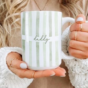 May include: White ceramic mug with a green striped pattern and the word "daddy" in a cursive font. The mug has a white handle and is held by a person wearing rings and a white sweater.