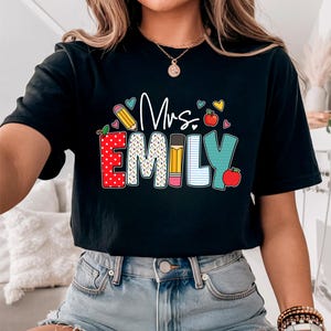 May include: Black t-shirt with the text "Mrs. Emily" in colorful letters with a school theme. The letters are decorated with hearts, pencils, and apples.