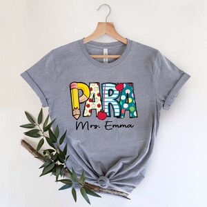 以下が含まれることがあります： グレーのTシャツに、鉛筆、リンゴ、その他の学校をテーマにしたデザインで「PARA」という言葉がカラフルに描かれたグラフィックがあります。「Mrs. Emma」というテキストがグラフィックの下にあります。