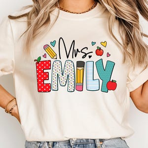 May include: A white t-shirt with the text "Mrs. Emily" in colorful letters with a pencil, apple, and heart designs.