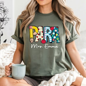 以下が含まれることがあります： カラフルな文字で「PARA」と書かれた緑色のTシャツ。各文字は鉛筆、リンゴ、ボタンなどの学校をテーマにしたパターンで飾られています。「Mrs. Emma」という文字が「PARA」の下に印刷されています。