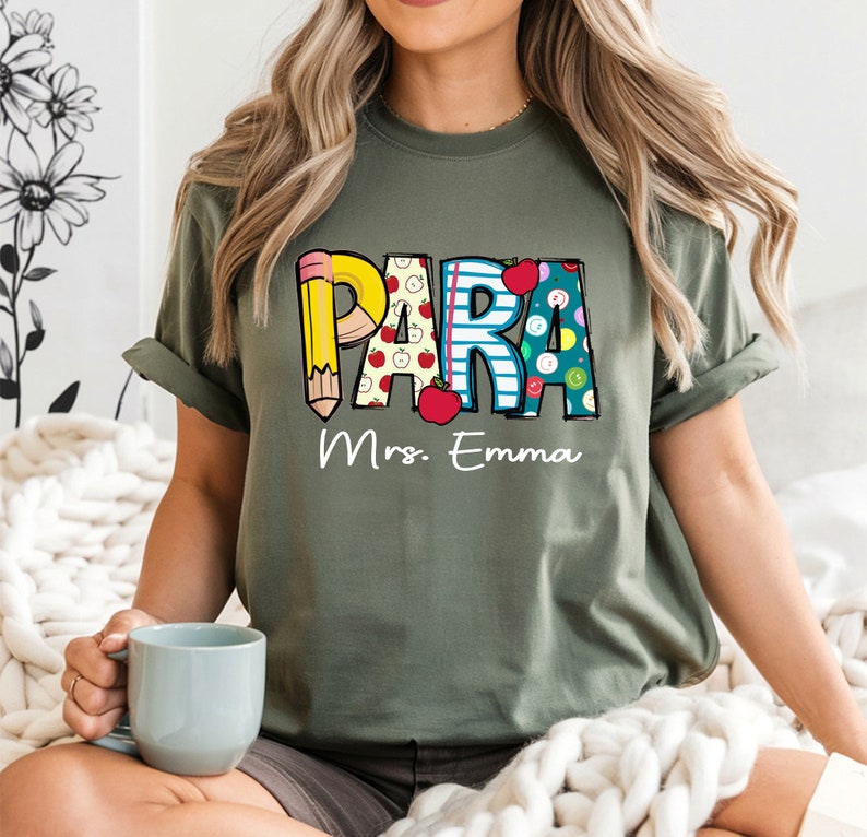 以下が含まれることがあります： カラフルな文字で「PARA」と書かれた緑色のTシャツ。各文字は鉛筆、リンゴ、ボタンなどの学校をテーマにしたパターンで飾られています。「Mrs. Emma」という文字が「PARA」の下に印刷されています。