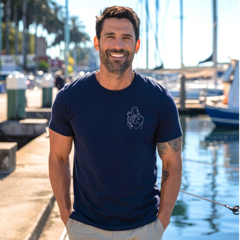May include: Navy blue t-shirt with a white line drawing of a person holding a baby. The t-shirt is short-sleeved and has a crew neck. The person in the image is smiling and wearing khaki pants.