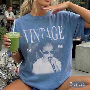 May include: A blue jean colored short-sleeve t-shirt featuring the white text VINTAGE EST 1986 above a graphic of a person wearing sunglasses and holding a microphone. The shirt has a crew neckline and a relaxed fit with a faded, washed-out appearance.
