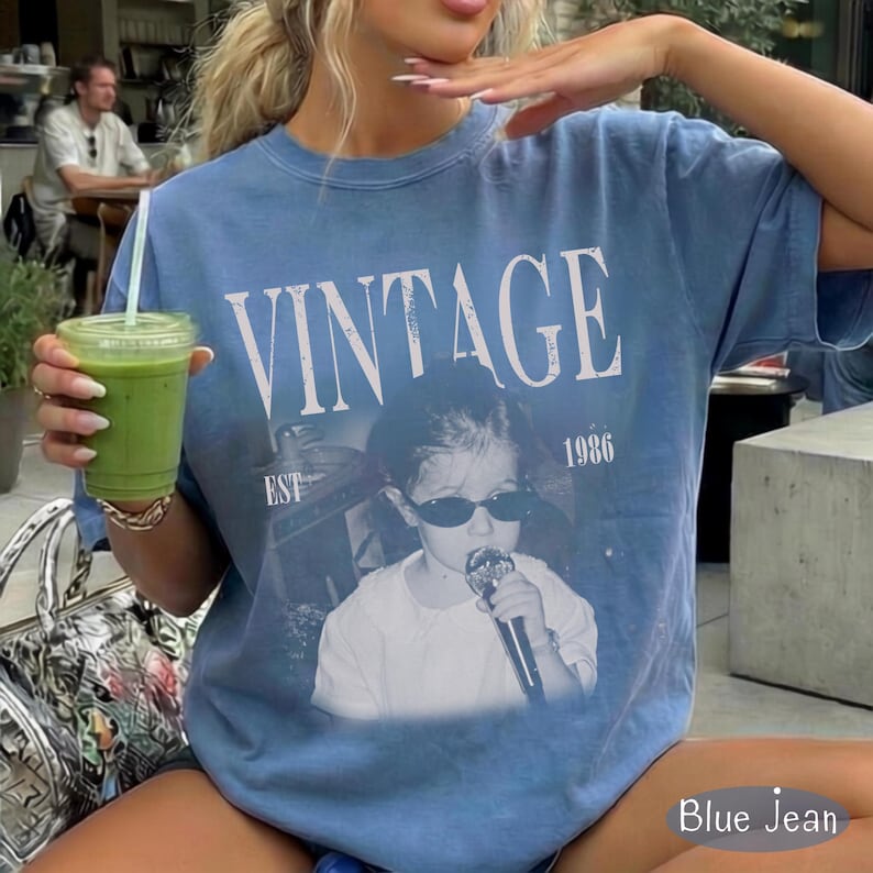 May include: A blue jean colored short-sleeve t-shirt featuring the white text VINTAGE EST 1986 above a graphic of a person wearing sunglasses and holding a microphone. The shirt has a crew neckline and a relaxed fit with a faded, washed-out appearance.