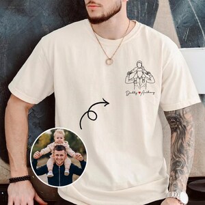 May include: Cream-colored t-shirt featuring a line drawing of a person carrying a child on their shoulders, with the text "Daddy Anthony" below. A photo of a person carrying a child on their shoulders is in a circle.