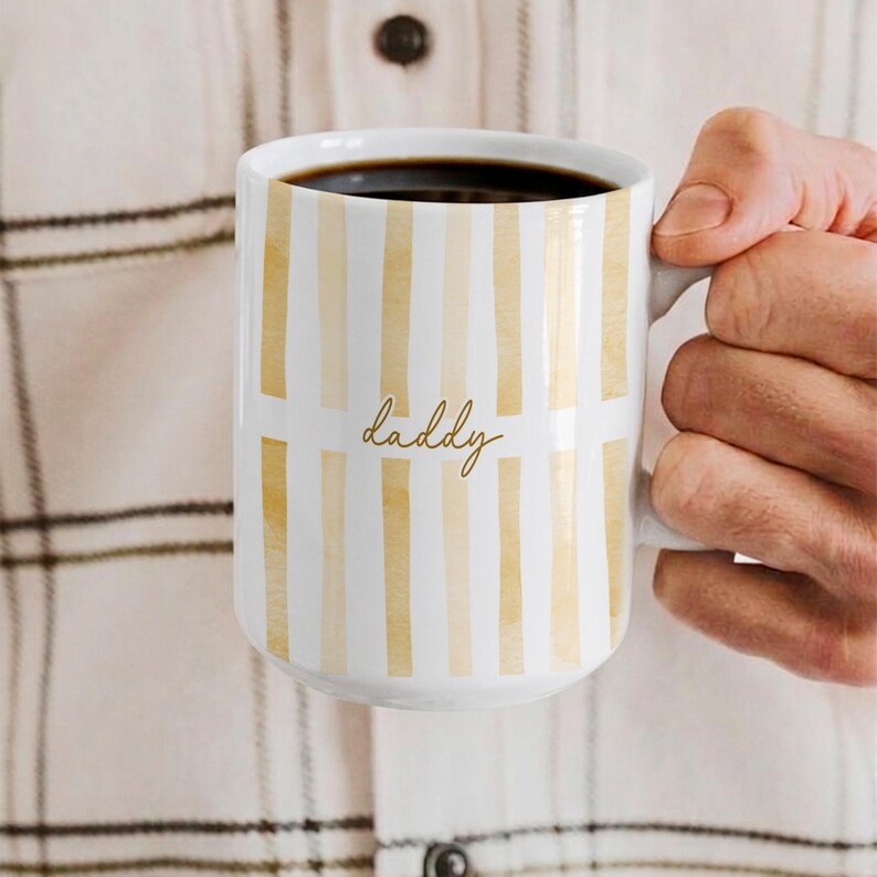 May include: White ceramic mug with vertical gold stripes and the word "daddy" in gold script. The mug is filled with dark coffee and has a large handle. A perfect gift for Father's Day.