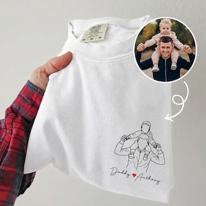 May include: White Comfort Colors t-shirt with a line drawing of a person carrying a child on their shoulders. The text "Daddy ❤️ Anthony" is below the drawing. A circular photo of a person with a child is in the upper right corner.