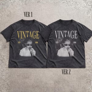 May include: Two charcoal gray short-sleeve t-shirts featuring a graphic print of a person wearing sunglasses and holding a microphone. The text VINTAGE, EST, and 1986 are printed on the front. The left shirt has gold text, while the right shirt has white text.