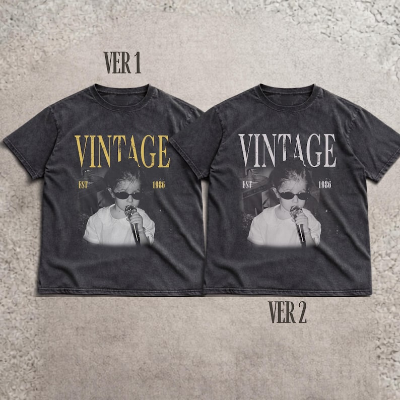 May include: Two charcoal gray short-sleeve t-shirts featuring a graphic print of a person wearing sunglasses and holding a microphone. The text VINTAGE, EST, and 1986 are printed on the front. The left shirt has gold text, while the right shirt has white text.