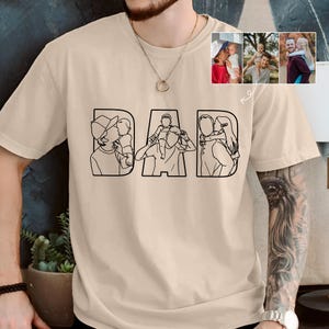 Peut inclure: T-shirt beige avec le mot "DAD" en lettres grasses et soulign&eacute;es. Chaque lettre pr&eacute;sente un dessin au trait noir d'un parent avec un enfant. De petites photos suppl&eacute;mentaires montrent des moments similaires parent-enfant. Un t-shirt d&eacute;contract&eacute;.
