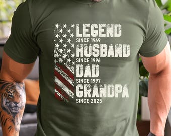 May include: Olive green t-shirt with a distressed American flag design. The shirt features the words "LEGEND SINCE 1969, HUSBAND SINCE 1996, DAD SINCE 1997, GRANDPA SINCE 2025" in white, distressed font.