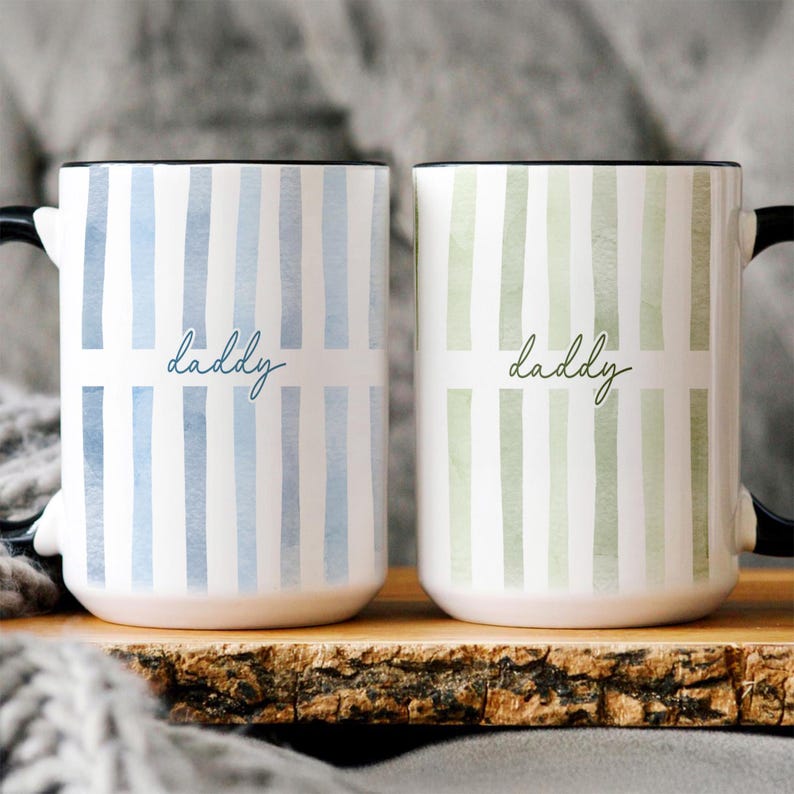 May include: Two white ceramic mugs with black handles and rims. One mug has blue stripes and the word "daddy" in script, the other has green stripes and the same text. The mugs are on a wooden surface.