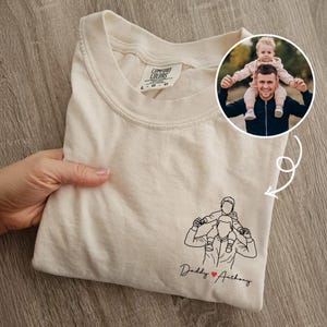 May include: Cream-colored t-shirt with a line drawing of a person carrying a child on their shoulders. The text "Daddy ❤️ Anthony" is below the drawing. A circular photo of a person carrying a child is in the upper right corner.