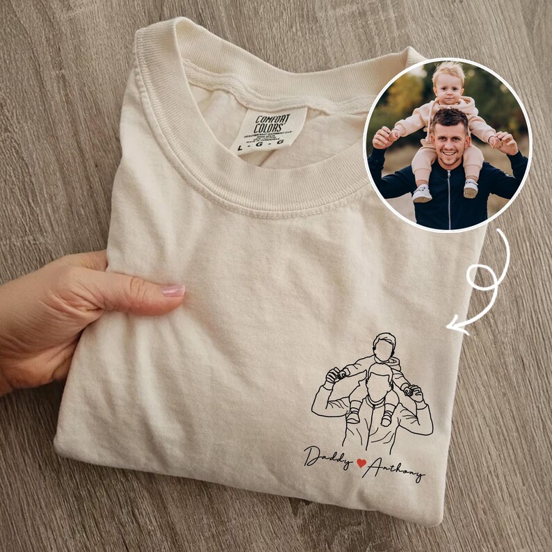May include: Cream-colored t-shirt with a line drawing of a person carrying a child on their shoulders. The text "Daddy ❤️ Anthony" is below the drawing. A circular photo of a person carrying a child is in the upper right corner.