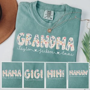 May include: A teal Comfort Colors t-shirt with the word "GRANDMA" in a floral design. Below the word are the names Taylor, Jackson, and Emma. The image also shows other shirts with the words "MAMA", "GIGI", "MIMI", and "MAMAW". A silver necklace is draped across the shirt.