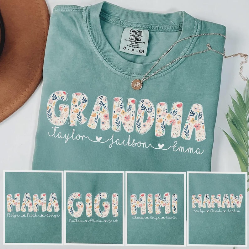 May include: A teal Comfort Colors t-shirt with the word "GRANDMA" in a floral design. Below the word are the names Taylor, Jackson, and Emma. The image also shows other shirts with the words "MAMA", "GIGI", "MIMI", and "MAMAW". A silver necklace is draped across the shirt.
