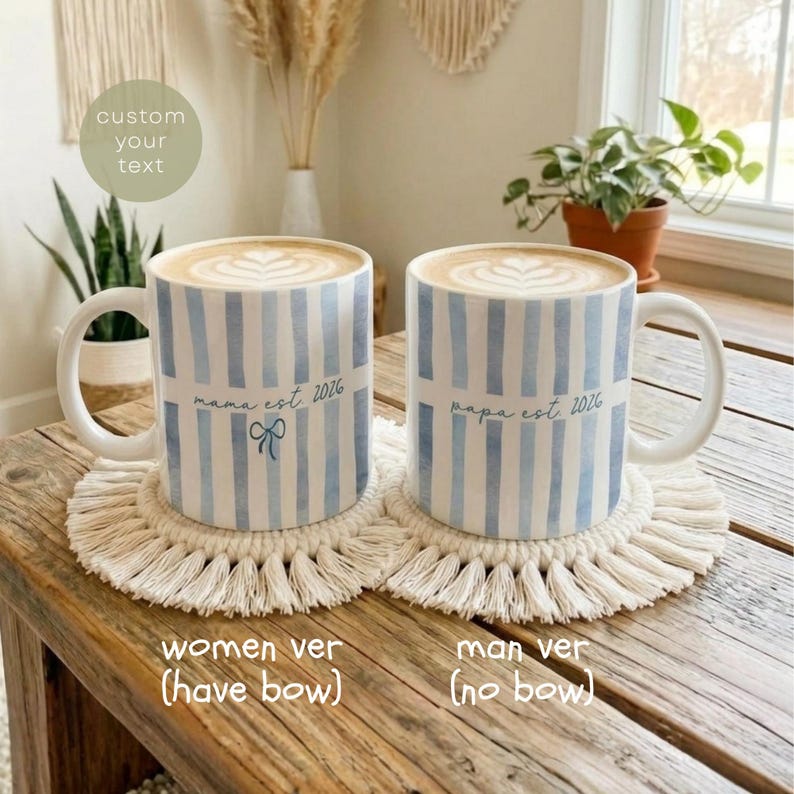 May include: Two white ceramic mugs with blue striped patterns, each filled with coffee and latte art, sit on macrame coasters. One mug says "mama est 2026" with a bow, the other says "papa est 2026".