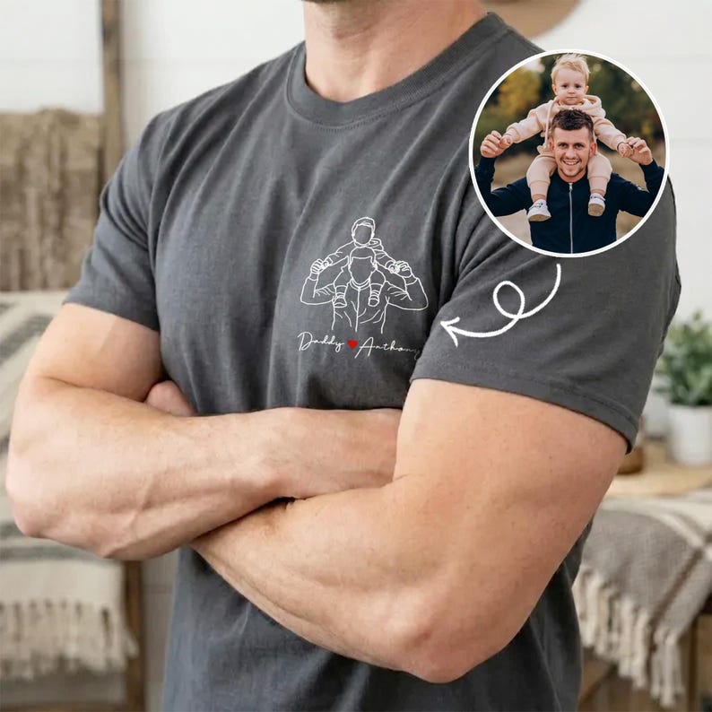 May include: Dark gray t-shirt featuring a line drawing of a person carrying a child on their shoulders, with the text "Daddy's Anchor" and a heart. A circular photo of a person and child is on the shirt.