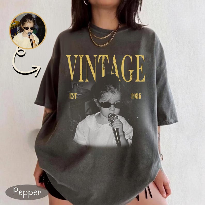 May include: A charcoal gray oversized t-shirt featuring a graphic print of a child wearing dark sunglasses and holding a microphone. The text VINTAGE EST 1986 is printed in yellow above and below the black and white photographic image.