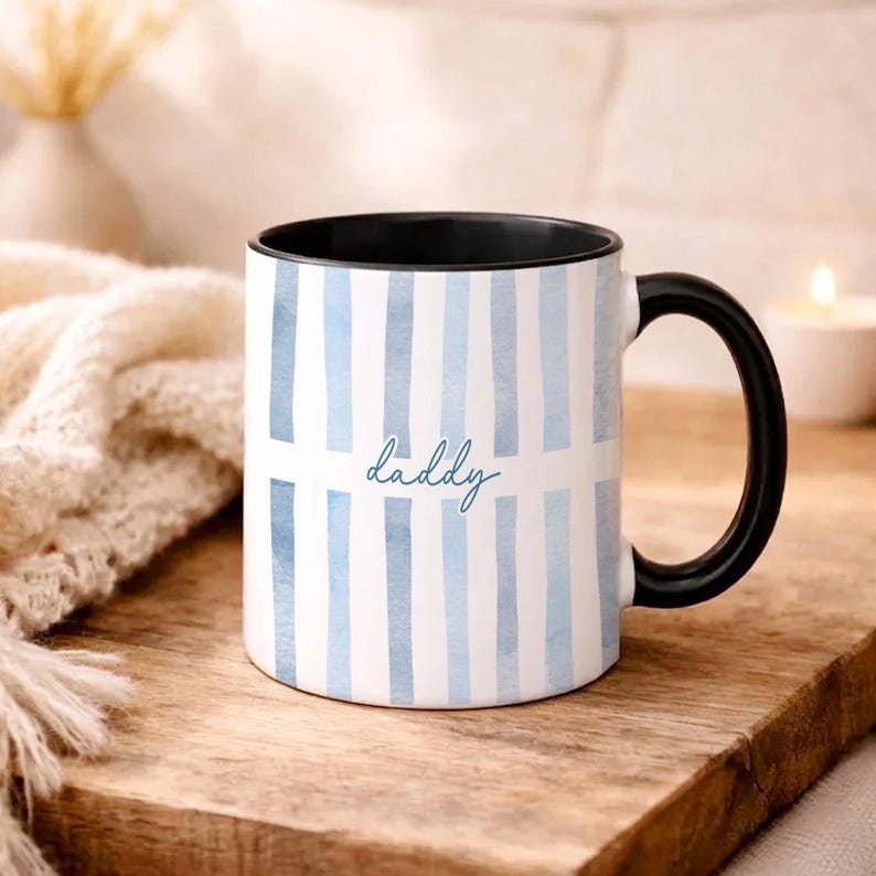 May include: White ceramic mug with blue watercolor vertical stripes and the word "daddy" in a script font. The mug has a black handle and interior. The mug is on a wooden surface.