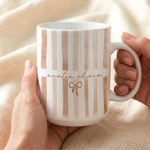 May include: White ceramic mug with vertical blush-colored stripes. The mug features the text "auntie olivia" in a delicate script font, along with a bow graphic. The mug is held by two hands.