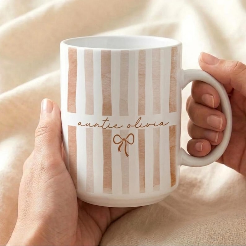 May include: White ceramic mug with vertical blush-colored stripes. The mug features the text "auntie olivia" in a delicate script font, along with a bow graphic. The mug is held by two hands.