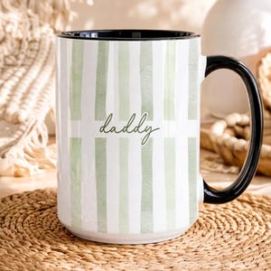May include: A white ceramic mug with a black handle and rim. The mug features a vertical striped pattern in light green and white, with the word "daddy" in a cursive font.