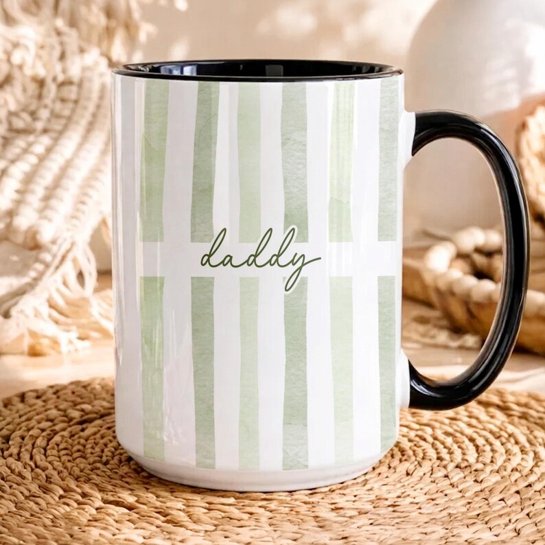 May include: A white ceramic mug with a black handle and rim. The mug features a vertical striped pattern in light green and white, with the word "daddy" in a cursive font.