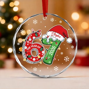 May include: A circular clear glass Christmas ornament featuring the number 67 in red and green with white polka dots. The number 6 is wrapped in colorful holiday lights, and the 7 wears a Santa hat with the name Skyler and the year 2026 printed.