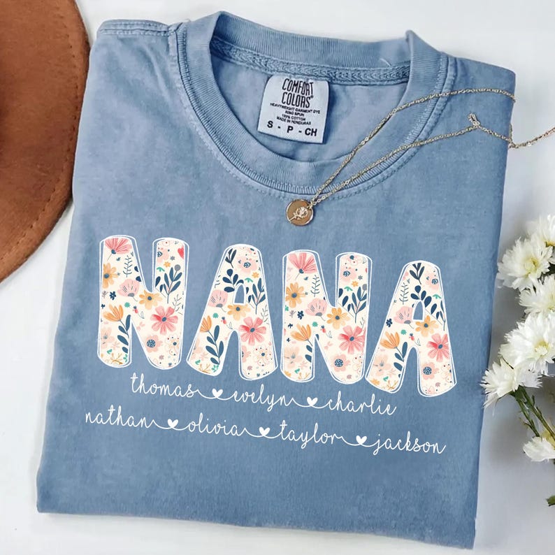 May include: A light blue Comfort Colors t-shirt with the word "NANA" in large floral letters. Below the word "NANA" are the names Thomas, Welyn, Charlie, Nathan, Olivia, Taylor, and Jackson. A gold necklace rests on the shirt.