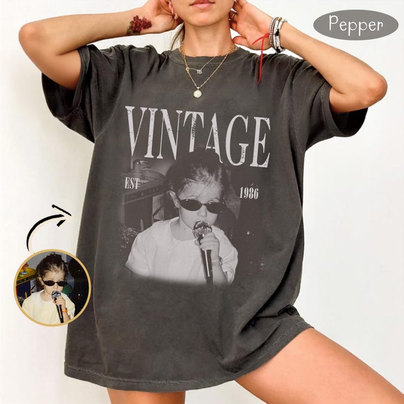 May include: A charcoal gray short-sleeve t-shirt featuring a graphic print of a person wearing sunglasses and holding a microphone. The text VINTAGE EST 1986 is printed above the image. The shirt has a crew neckline and a relaxed, loose fit.
