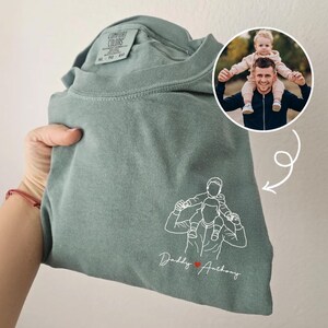 May include: Sage green Comfort Colors t-shirt with a white line drawing of a person carrying a child on their shoulders. The text "Daddy Anthony" is below the drawing, with a red heart. A photo of a man and child is in a circle.