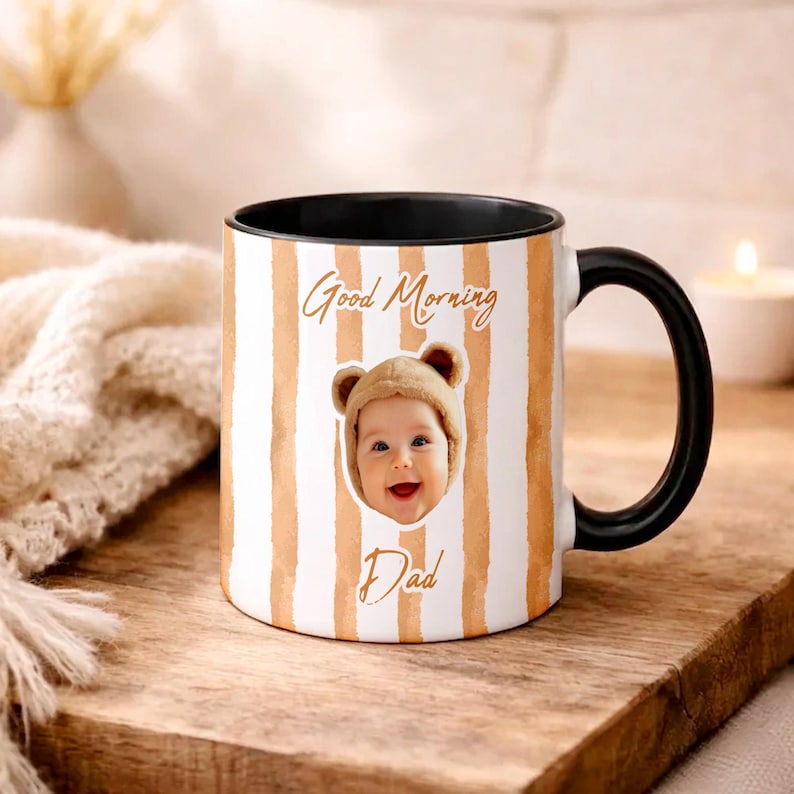 May include: A white ceramic mug with a black handle and interior, featuring vertical tan watercolor stripes. The mug displays a photograph of a baby wearing a bear-ear hat, with the text Good Morning printed above and the word Dad printed below.