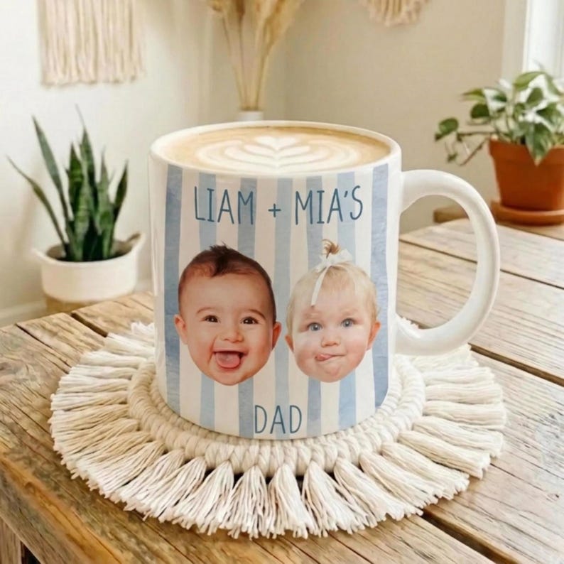 May include: White ceramic mug with a handle, featuring a blue and white striped design. The mug has two baby faces and the text "LIAM + MIA'S DAD". The mug is on a macrame coaster, with a coffee drink inside.