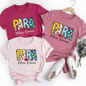 以下が含まれることがあります： ピンクとマルーンのTシャツ3枚に、カラフルな文字で「PARA」と書いてあり、それぞれの下に「Mrs. Emma」と印刷されています。シャツは折り畳んで、白いスニーカーと緑の葉っぱと一緒に白い背景の上に置かれています。