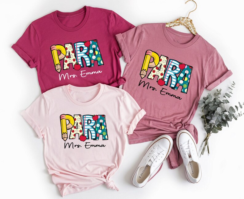 以下が含まれることがあります： ピンクとマルーンのTシャツ3枚に、カラフルな文字で「PARA」と書いてあり、それぞれの下に「Mrs. Emma」と印刷されています。シャツは折り畳んで、白いスニーカーと緑の葉っぱと一緒に白い背景の上に置かれています。