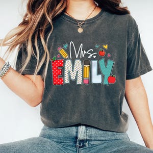 May include: A dark gray t-shirt with the text "Mrs. Emily" in colorful letters. The letters are decorated with images of apples, pencils, and notebook paper.