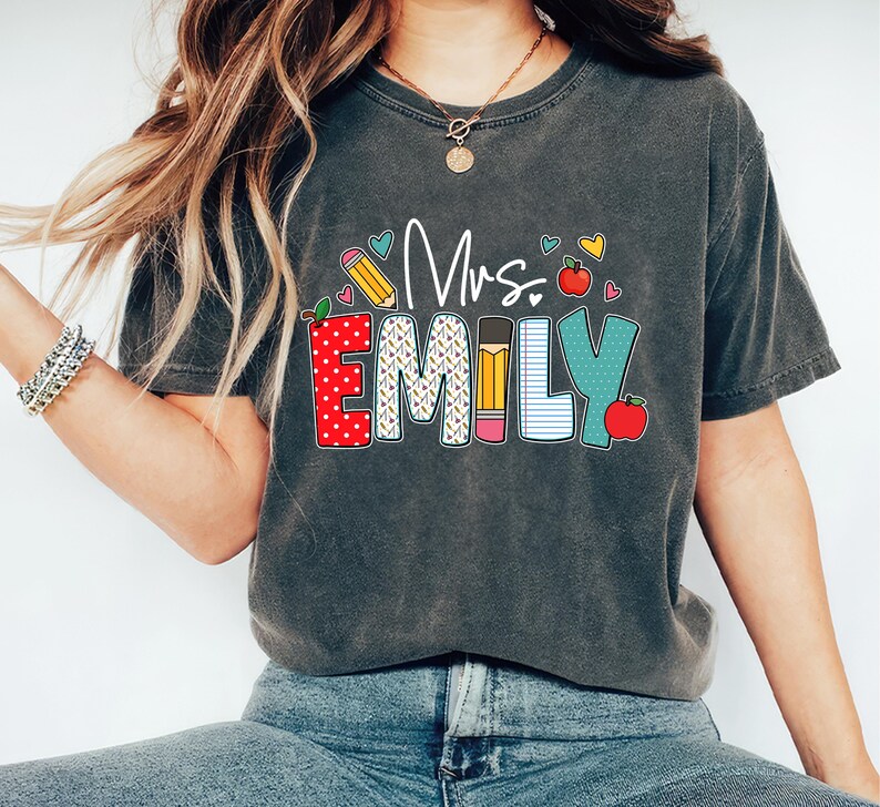 May include: A dark gray t-shirt with the text "Mrs. Emily" in colorful letters. The letters are decorated with images of apples, pencils, and notebook paper.