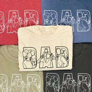 Peut inclure: Plusieurs t-shirts de couleurs vari&eacute;es, dont rouge, bleu et beige. Chaque t-shirt affiche le mot "DAD" en blanc, avec un dessin au trait diff&eacute;rent d'un parent et d'un enfant dans chaque lettre.