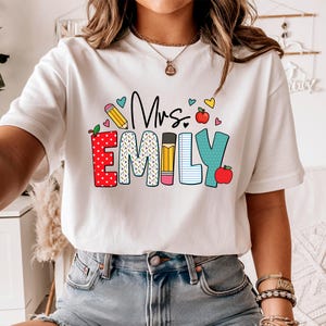 May include: White t-shirt with a colorful graphic that says "Mrs. Emily" with a red apple, pencil, and hearts.