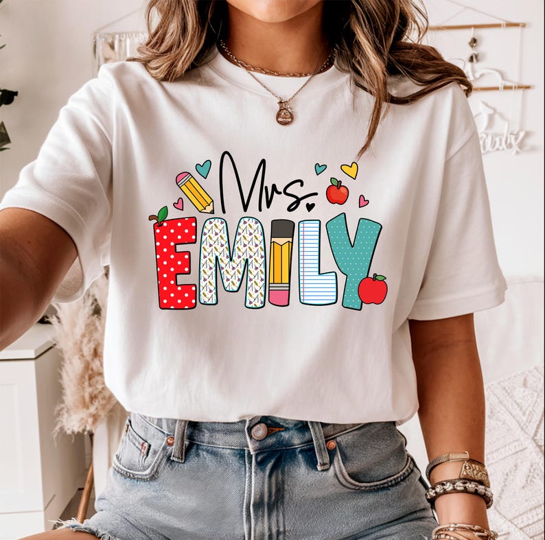May include: White t-shirt with a colorful graphic that says "Mrs. Emily" with a red apple, pencil, and hearts.