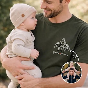 May include: An olive green t-shirt featuring a line drawing of a father carrying a child on his shoulders, with the text "Daddy Anthony". A baby in a beige outfit and hat is held by a man. A small photo of a father and child is in the lower right corner.