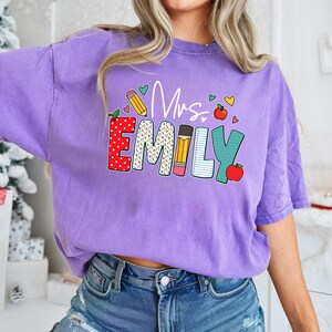 May include: Purple t-shirt with the text "Mrs. Emily" in colorful letters decorated with school supplies like pencils, an apple, and a notebook.