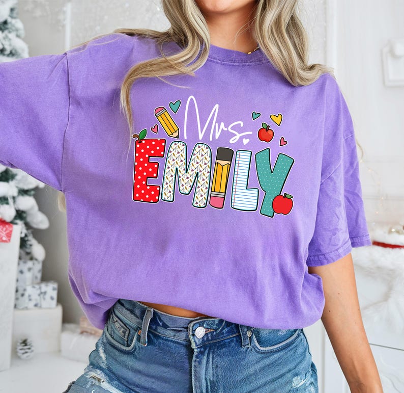 May include: Purple t-shirt with the text "Mrs. Emily" in colorful letters decorated with school supplies like pencils, an apple, and a notebook.