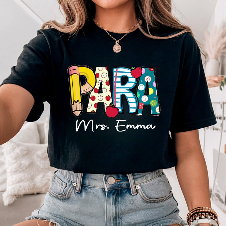 以下が含まれることがあります： カラフルなグラフィックがプリントされた黒いTシャツ。グラフィックには「PARA」の文字と、鉛筆、リンゴ、その他の学校をテーマにしたデザインが描かれています。「Mrs. Emma」という文字がグラフィックの下にあります。