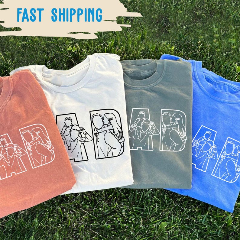 Peut inclure: Quatre t-shirts &agrave; manches courtes, corail, blanc, vert sauge et bleu, sont pr&eacute;sent&eacute;s sur de l'herbe verte. Chaque t-shirt affiche le mot "DAD" en blanc, avec un dessin au trait d'un parent et d'un enfant.