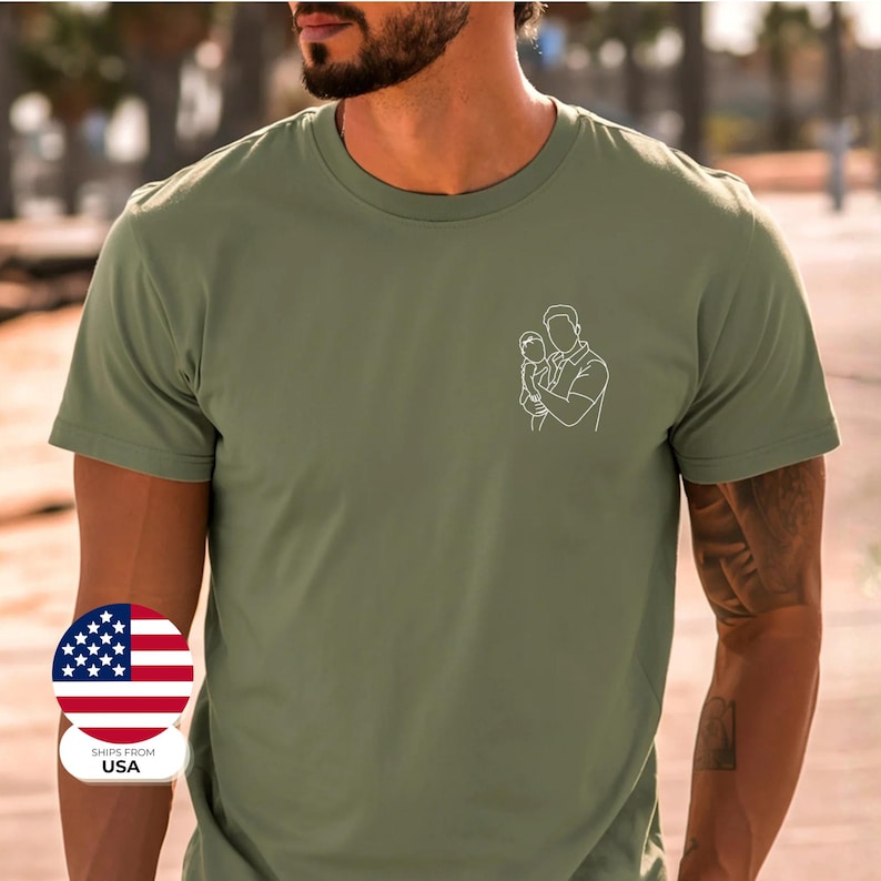 May include: Olive green t-shirt with a white line drawing of a parent holding a child on the left chest. The shirt has a crew neck and short sleeves. A small USA flag sticker is on the bottom left.