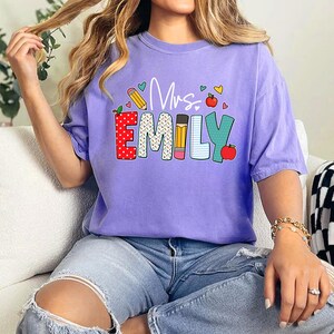 May include: Purple t-shirt with the text "Mrs. Emily" in colorful letters with a pencil, apple, and hearts design.